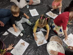 dzieci siedzą na rozłożonej na podłodze folii i malują bałwanki na kratkach papieru