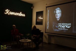 Pod neonem "Kameralna" siedzą Hanna Grabowska-Macioszek i Zbigniew Stryj. W tle ekran z wyświetloną okładką "Nie kocham teatru".
