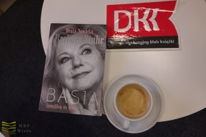 Na białym stoliku: banerek "DKK", książka "BASIA" i filiżanka kawy.