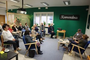 Autor rozmawia z dyrektorką biblioteki w salce kameralnej. Uczestnicy siedzą w około na fotelach i sofie. Na ciemnozielonej ścianie świeci neon "Kameralna".