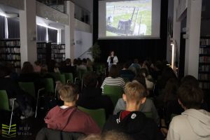 Prowadząca spotkanie w dużej sali biblioteki. Zdjęcie na dużym ekranie. Pełna widownia.