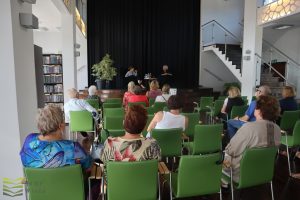 Widok w kierunku sceny z końca sali. Uczestnicy siedzą na zielonych krzesłach.